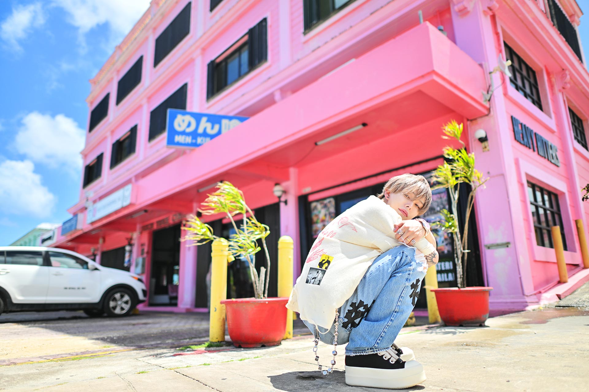 ピンクの建物前でしゃがみ込む華神疾翔、ポップな街並みとラフなスタイルが印象的な海外グラビア