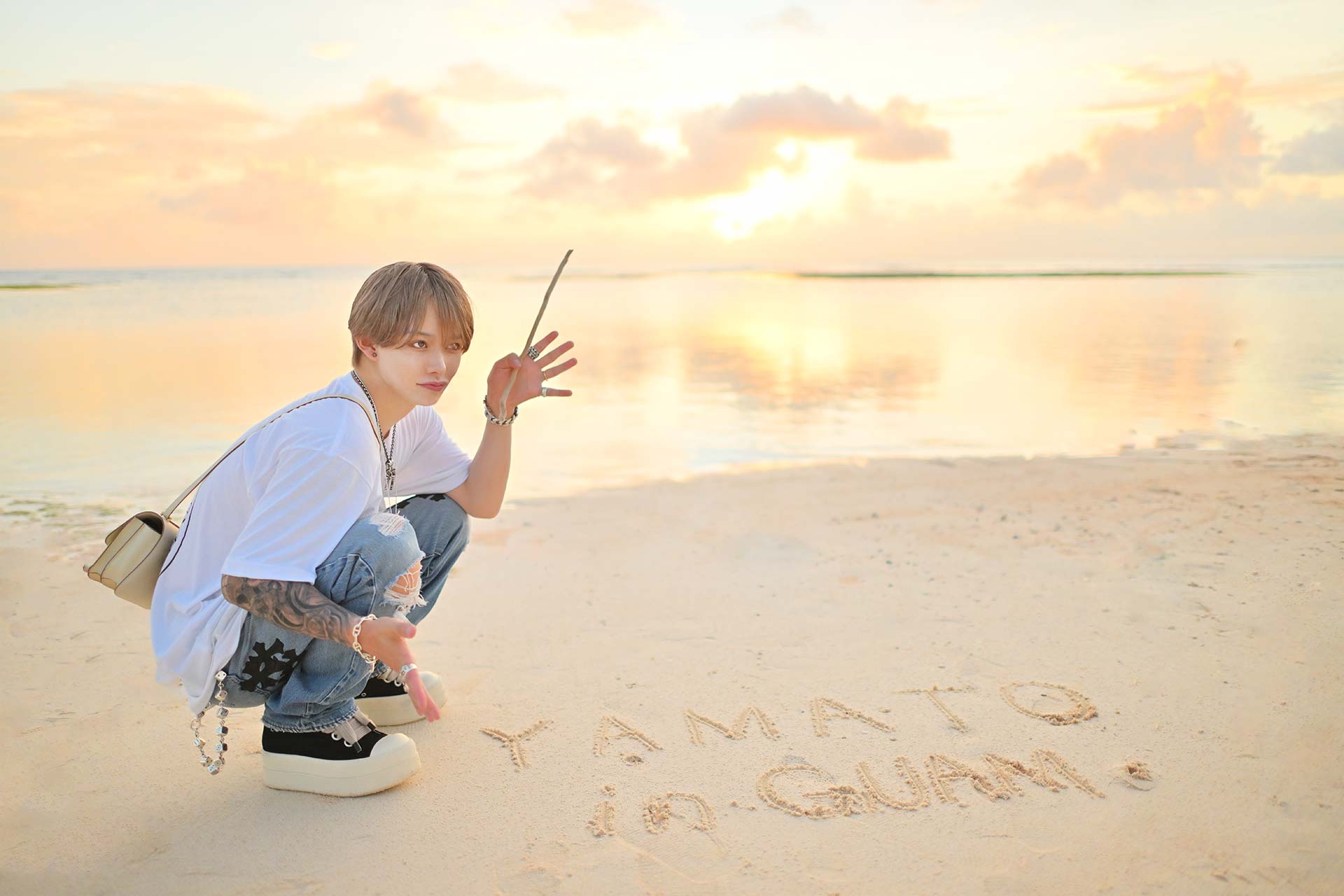 グアムの夕暮れビーチで砂浜にしゃがむ華神疾翔、穏やかな海と夕焼けが美しいリゾートショット