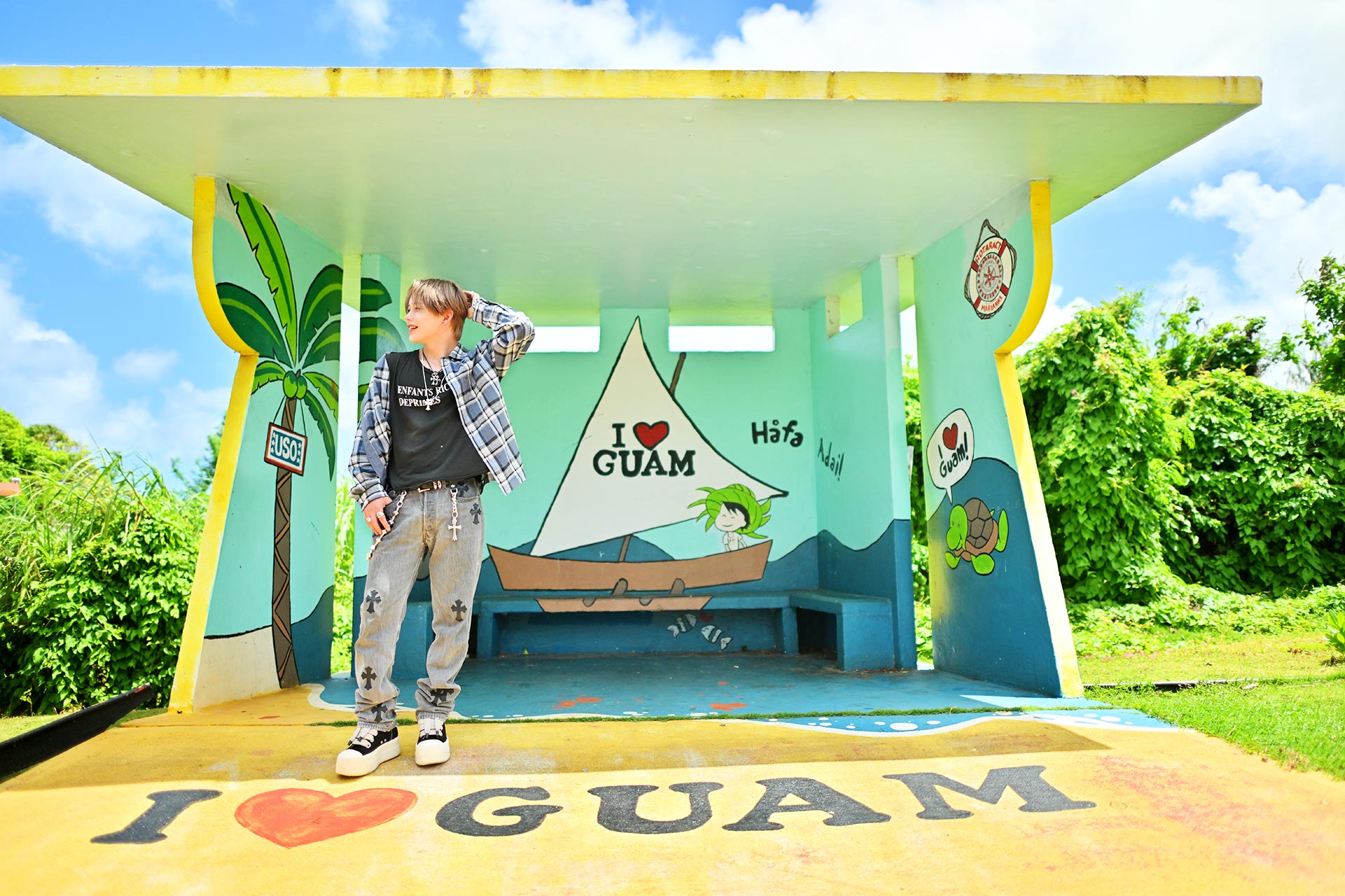 グアムの「I LOVE GUAM」スポット前で立ち止まり、華神疾翔が南国らしい爽やかな笑顔を見せる全身ショット。青空とヤシの木アートが映えるグアム旅行グラビア写真