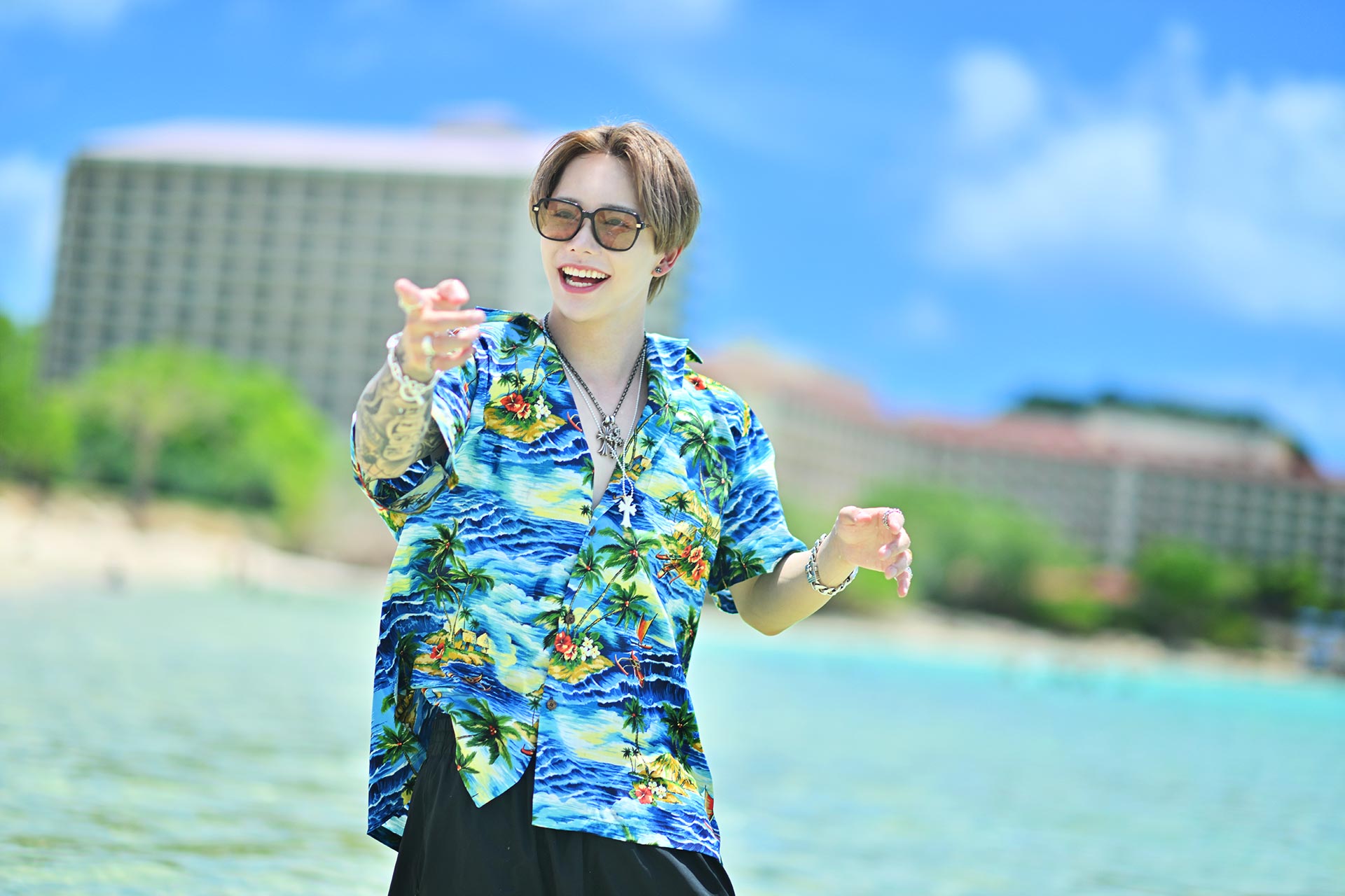 グアムの海辺で笑顔を見せながら手を差し伸べる華神疾翔。アロハシャツとエメラルドブルーの海が印象的なビーチ撮影カット